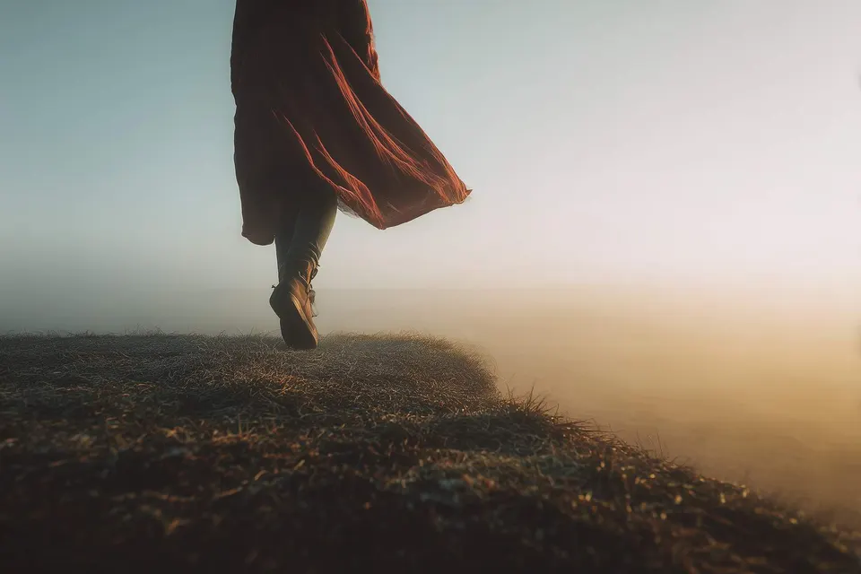 A silhouette in a red skirt walks toward a hazy horizon at the edge of a grassy cliff